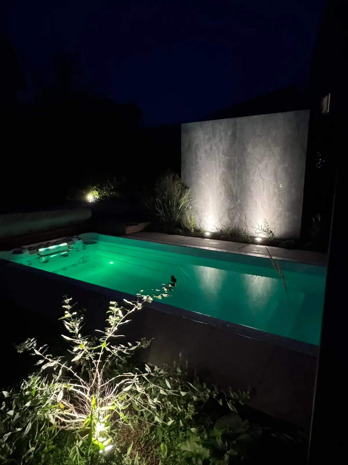 Swim spa INFINITAS completamente interrata davanti a un muro in pietra di notte